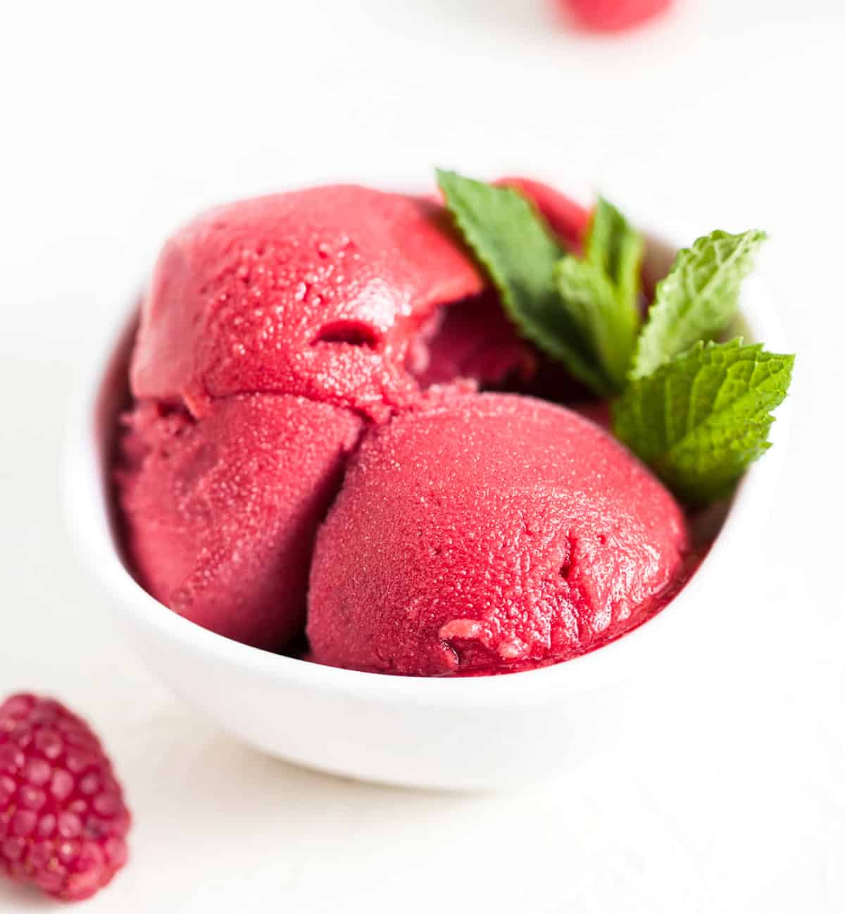 Scoops of raspberry sorbet in a white bowl with mint leaves on the side and a raspberry sitting in front of the bowl.