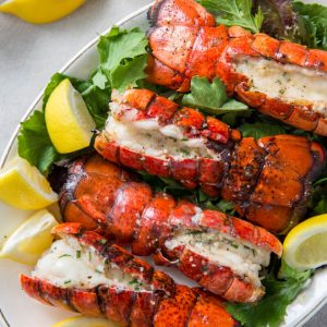 Four grilled lobster tails sitting on a plate with lemon wedges.