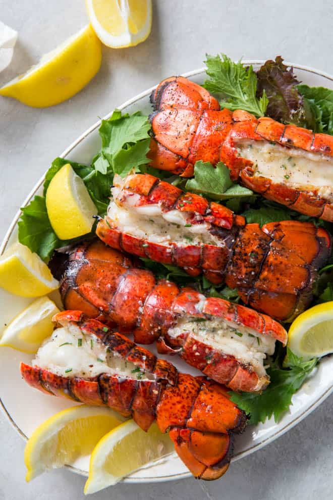 Four grilled lobster tails sitting on a plate with lemon wedges.