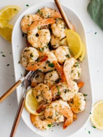 Cooked lemon garlic shrimp on a white serving plate with serving fork and lemon wedges.