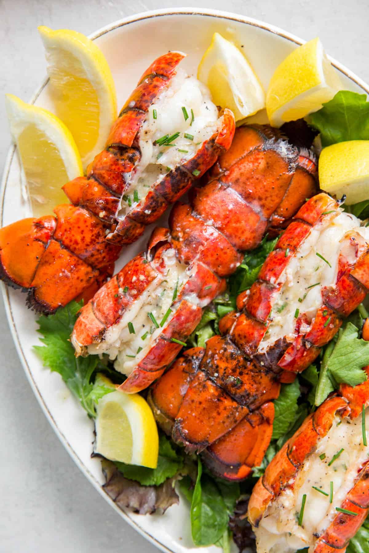 Four grilled lobster tails on a bed of lettuce on a plate with lemon wedges.