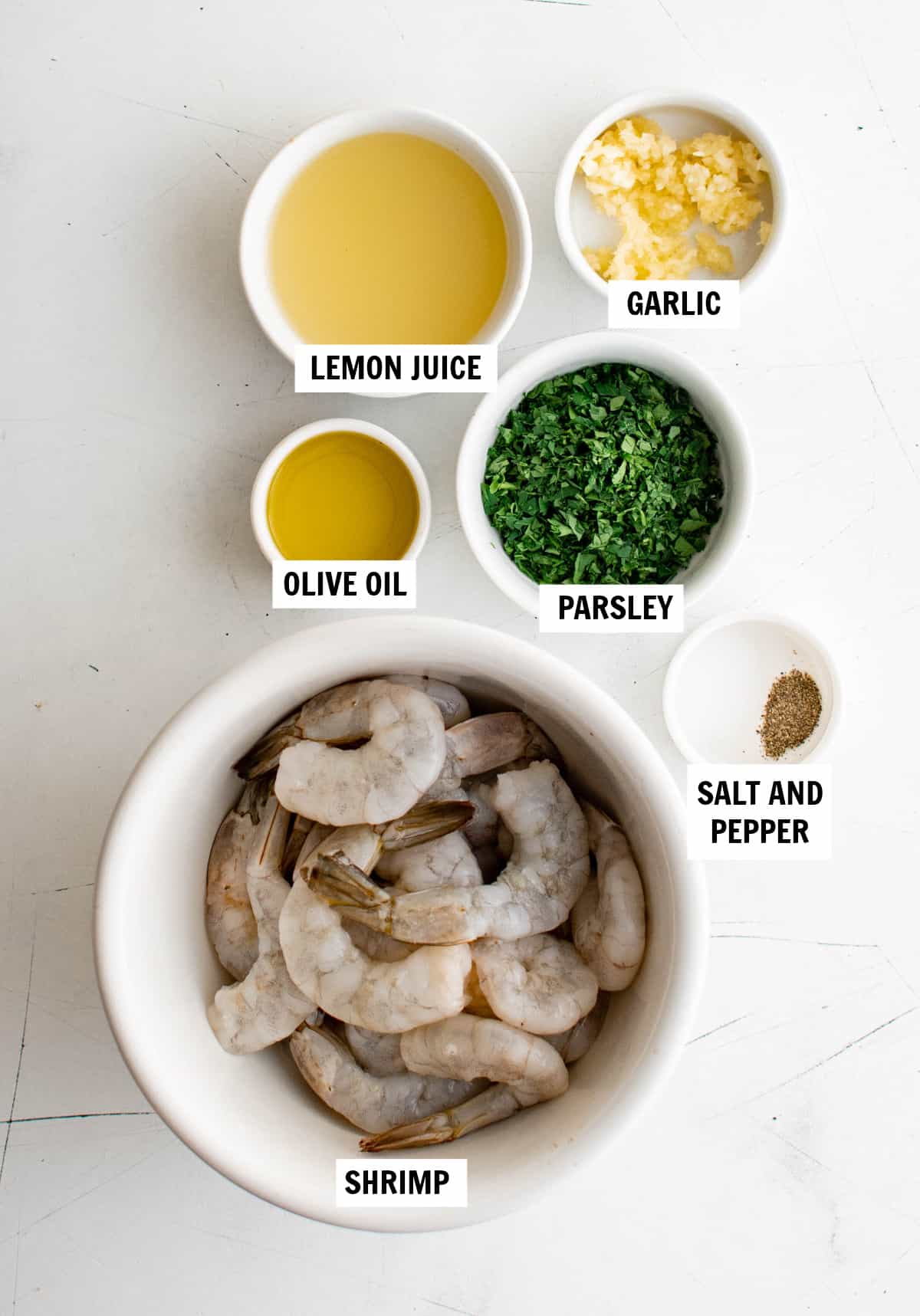 All of the ingredients for lemon garlic shrimp on a white countertop including peeled and deveined shrimp, olive oil, lemon juice, garlic, parsley, salt and pepper.