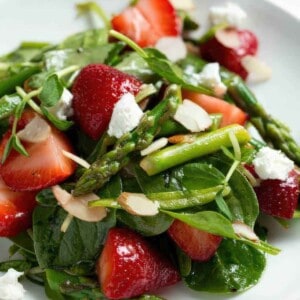 Strawberry spinach and asparagus salad topped with sliced almonds and goat cheese on a white plate.