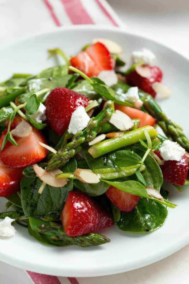 Strawberry spinach and asparagus salad topped with sliced almonds and goat cheese on a white plate.