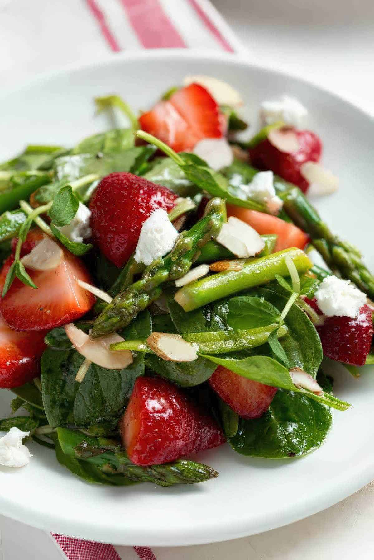 Strawberry spinach and asparagus salad topped with sliced almonds and goat cheese on a white plate.