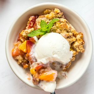 A large bowl of peach crumble with a scoop of vanilla ice cream and a spoon.