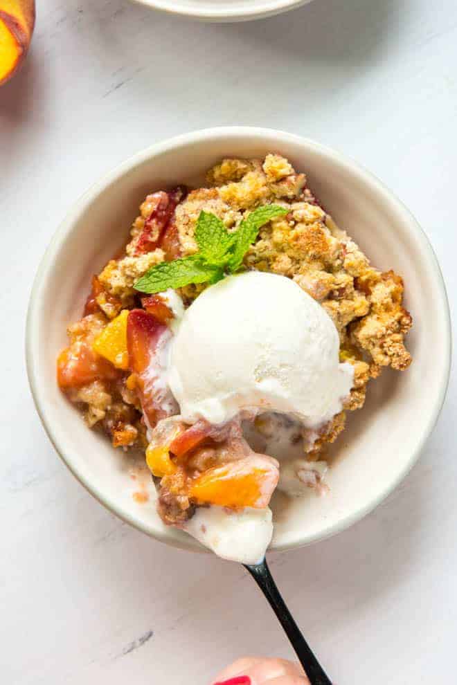 A large bowl of peach crumble with a scoop of vanilla ice cream and a spoon.