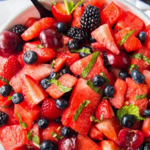 A large white serving bowl filled with watermelon, strawberries, blueberries, blackberries and cherries with a simple honey lemon dressing and mint. A large serving spoon rests in the bowl with the salad.
