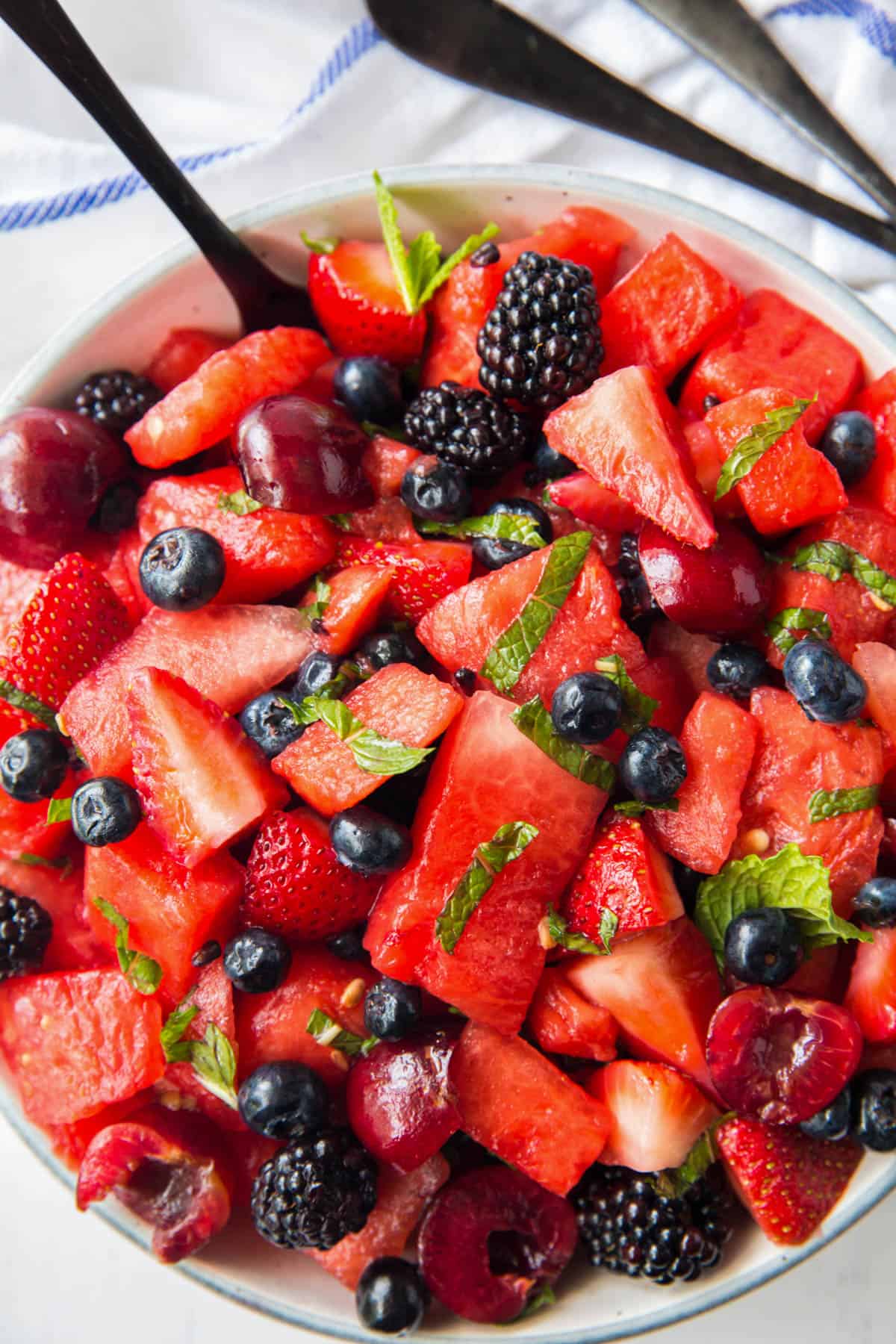 A large white serving bowl filled with watermelon, strawberries, blueberries, blackberries and cherries with a simple honey lemon dressing and mint. A large serving spoon rests in the bowl with the salad.