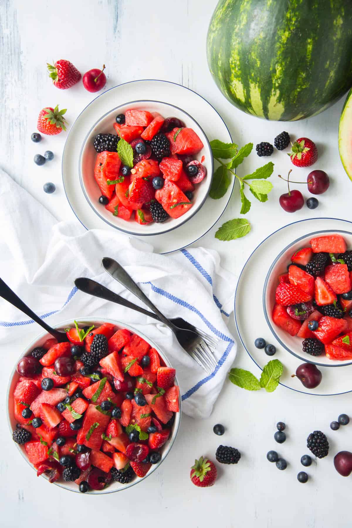 Two bowls with watermelon fruit salad sitting next to a large bowl with watermelon fruit salad. Forks sit on the side of the bowls.