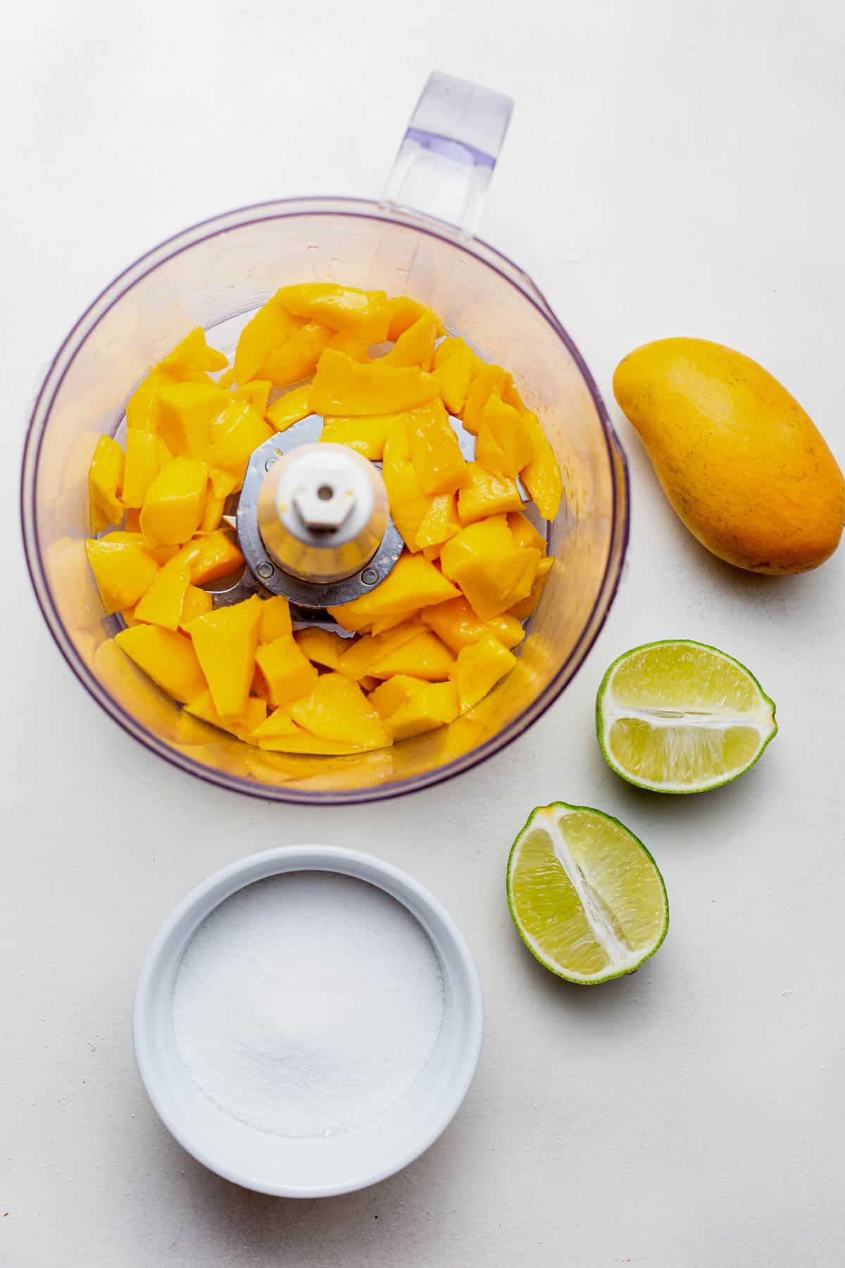 Fresh mango, limes and sugar on a white countertop.