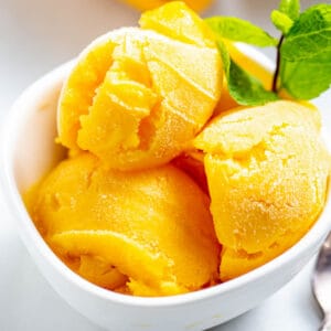 Three scoops of homemade mango sorbet in a white bowl with a sprig of peppermint on top.