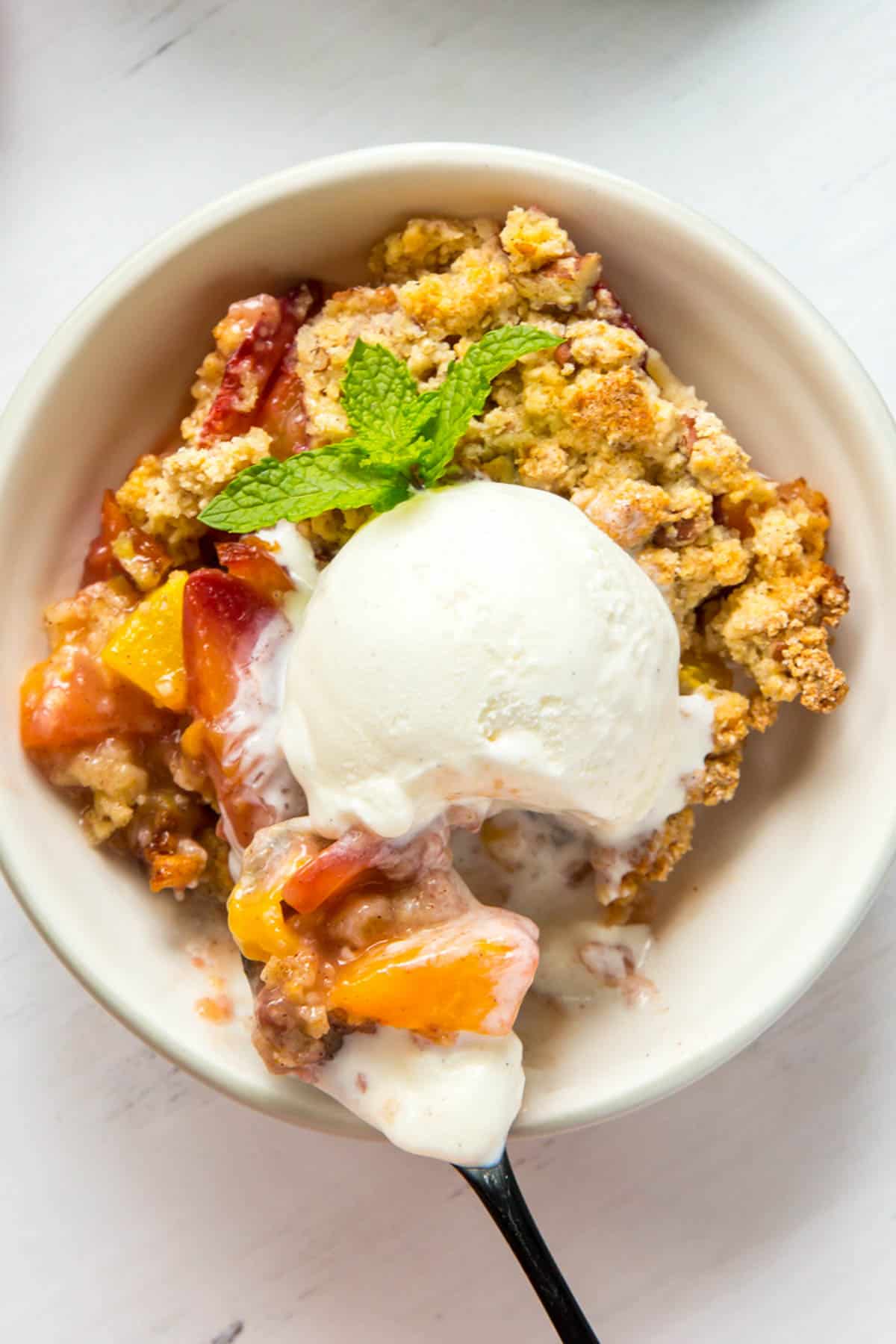 a white bowl filled with peach crumble and a scoop of vanilla ice cream