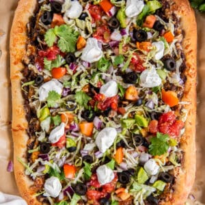 Homemade Taco Pizza on a piece of parchment paper on a baking sheet. The pizza is topped with chopped lettuce, tomatoes, green onions, black olives, salsa, cilantro and sour cream.