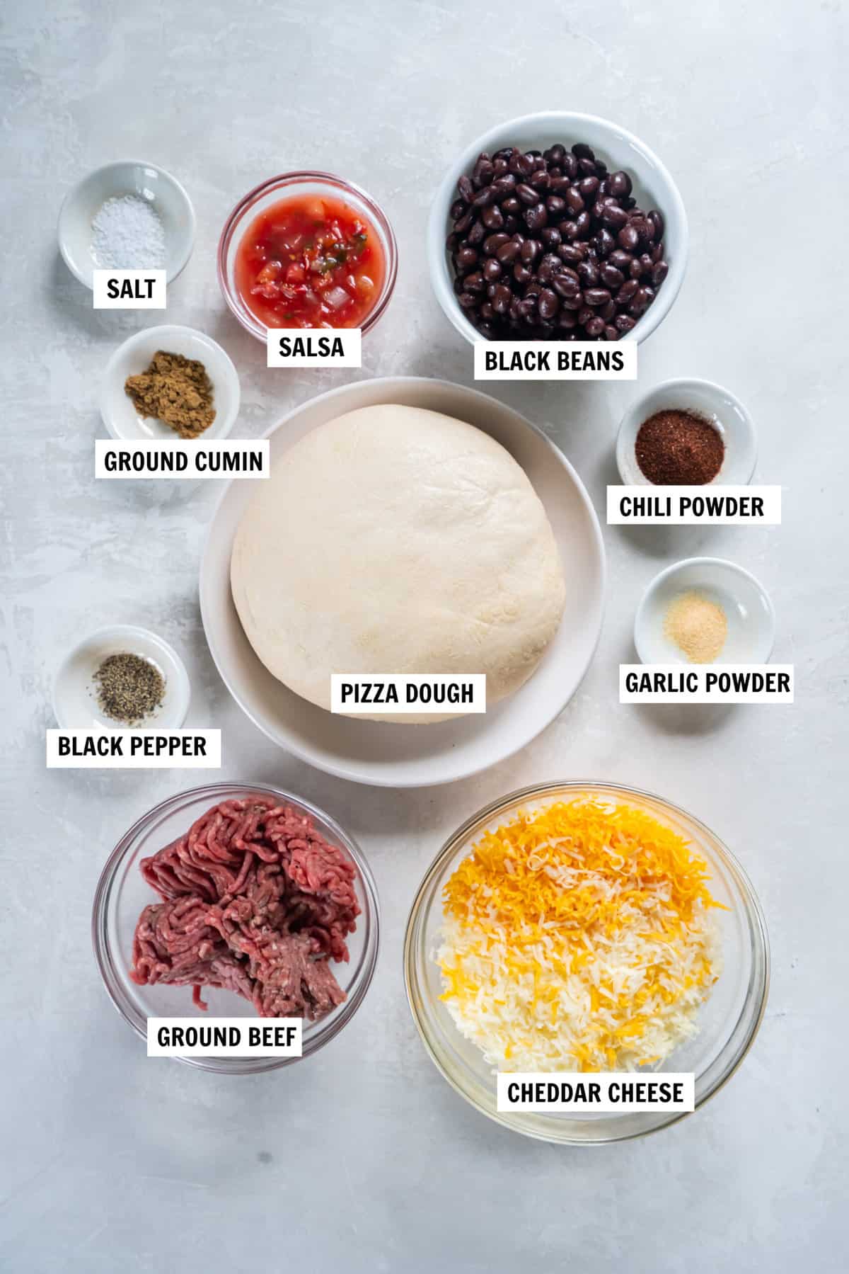 All of the ingredients for taco pizza on a white countertop in bowls including ground beef, chili powder, ground cumin, garlic powder, salt and pepper, black beans, salsa, chili powder, ground cumin, pizza dough and cheese.