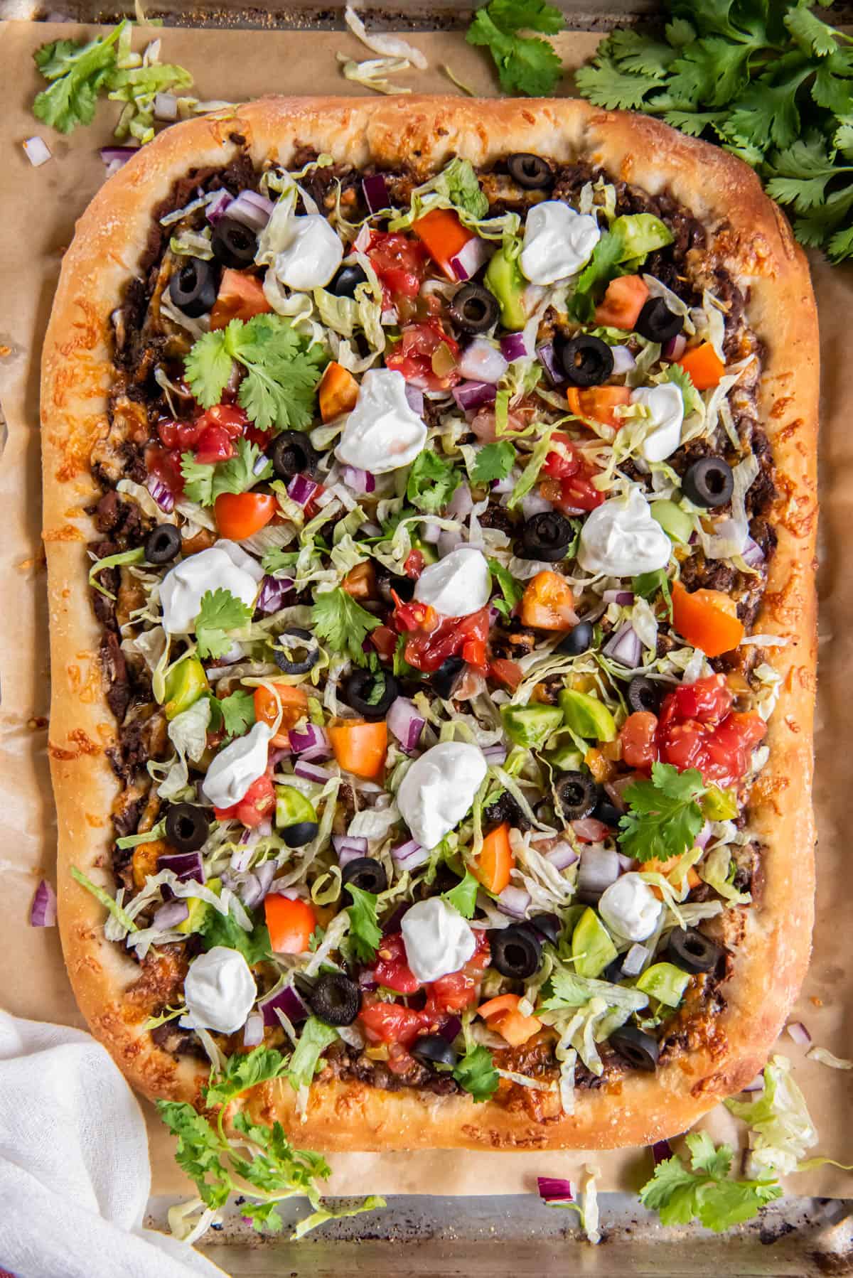 Homemade Taco Pizza on a piece of parchment paper on a baking sheet. The pizza is topped with chopped lettuce, tomatoes, green onions, black olives, salsa, cilantro and sour cream.