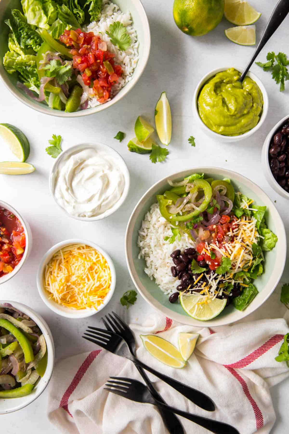 Two veggie burrito bowls sitting on a white countertop with small bowls of additional toppings including guacamole, sour cream, cheese and salsa.