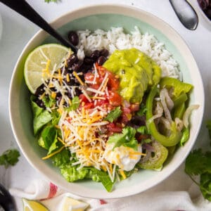 A white bowl filled with white rice, sauteed peppers and onions, guacamole, black beans, lettuce, pico de gallo and shredded cheese. A fork sits in the bowl.