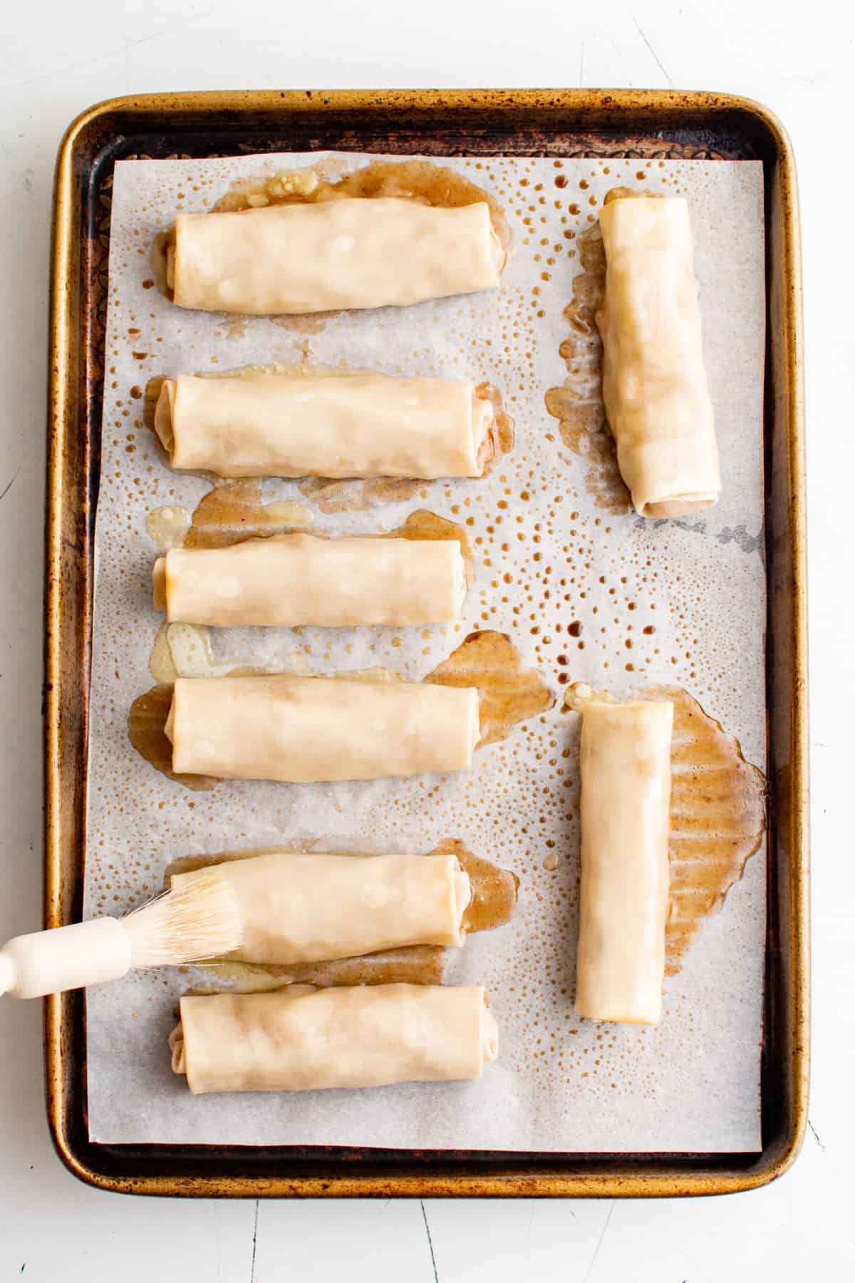 The baked egg rolls are on a sheet pan and a pastry brush is brushing them with butter before sprinkling them with cinnamon sugar.