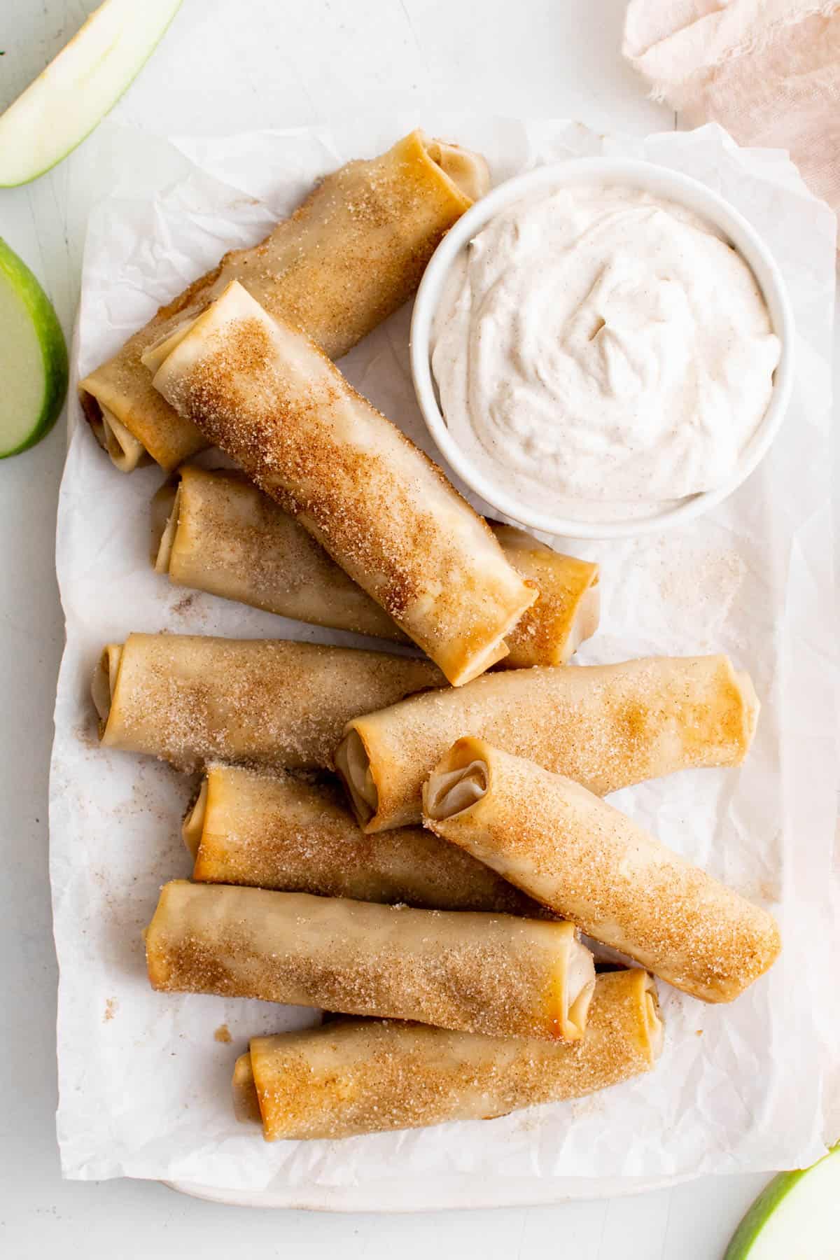 All of the baked apple pie egg rolls on a piece of parchment paper. The cinnamon whipped cream is sitting in a bowl next to the egg rolls.