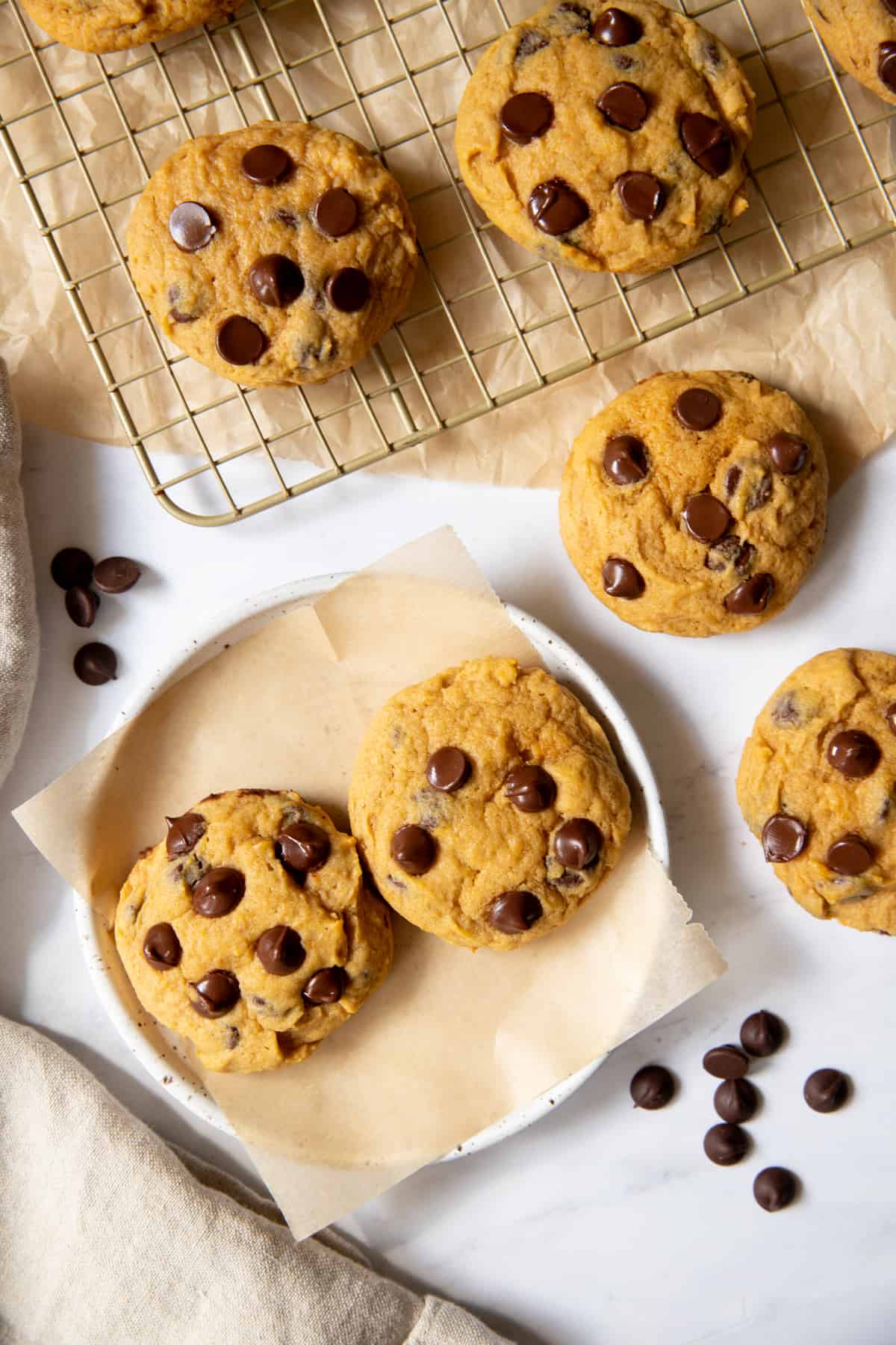 two pumpkin chocolate chip cookies on a plate next to a baking rack with more cookies
