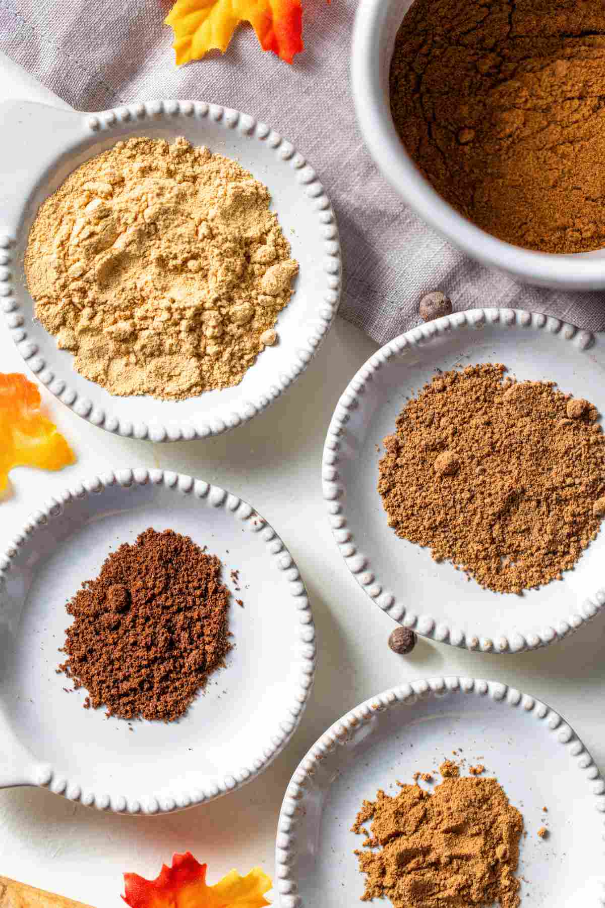The individual ground spices for pumpkin pie spice, cinnamon, ginger, nutmeg, allspice, and cloves, in small white dishes before mixing