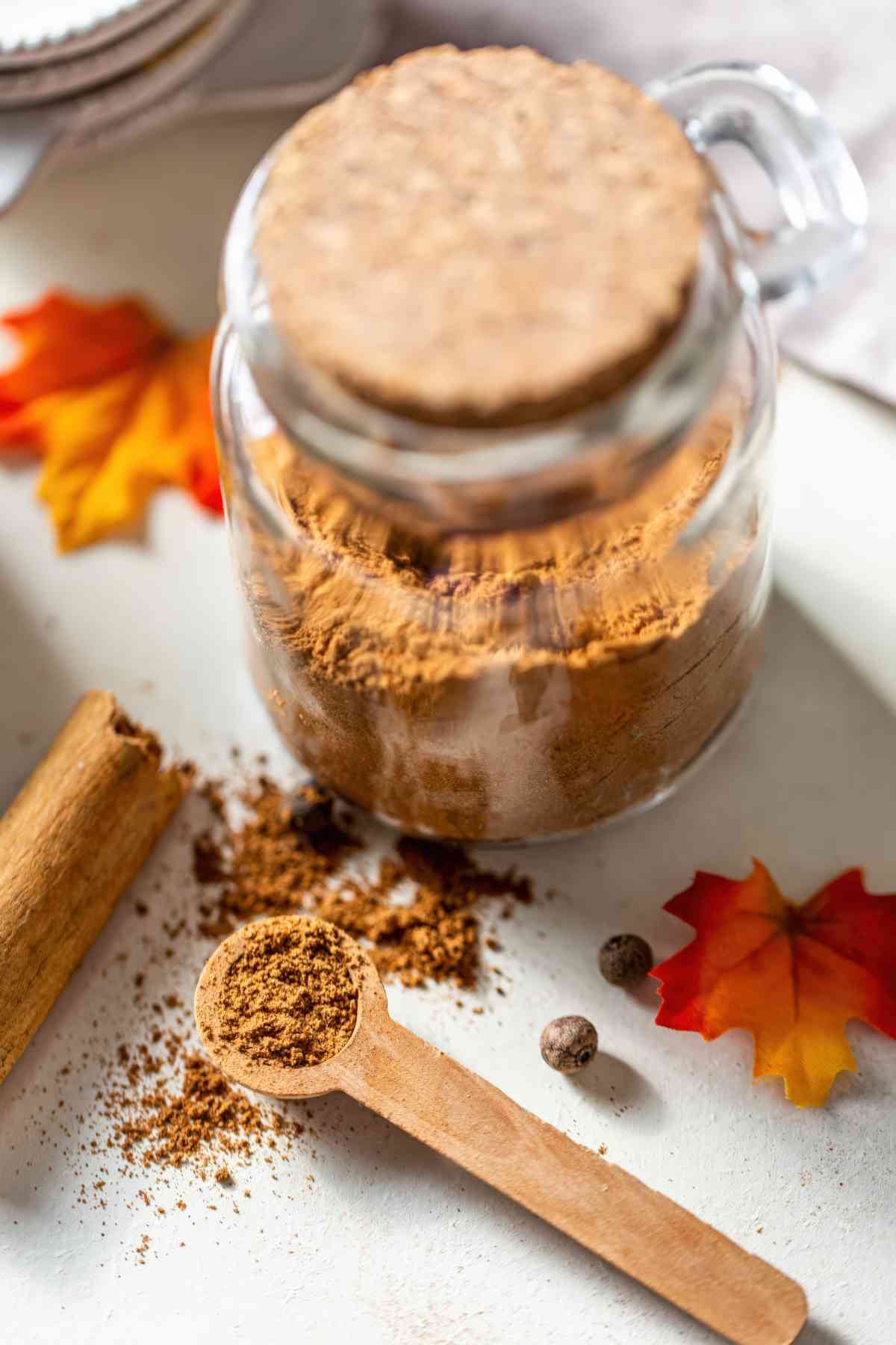 A measuring spoon of homemade pumpkin pie spice beside a filled jar, ready to use as a substitute for store-bought