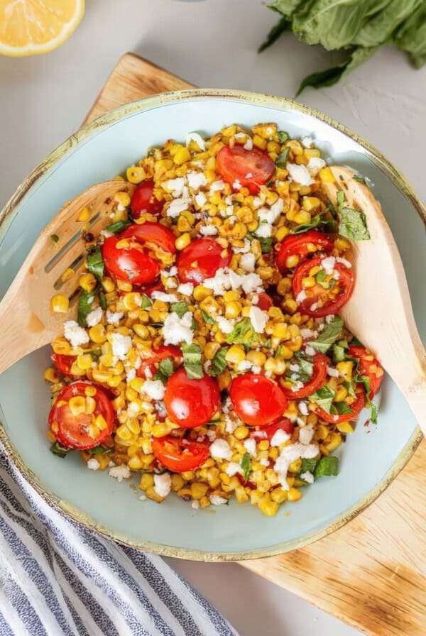 Overhead view of corn and tomato salad mixed with feta and basil, served in a light blue bowl with wooden utensils resting inside.