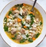 A bowl of soup with visible mini meatballs, spinach, carrots, and pasta, garnished with Parmesan flakes and served with a spoon.