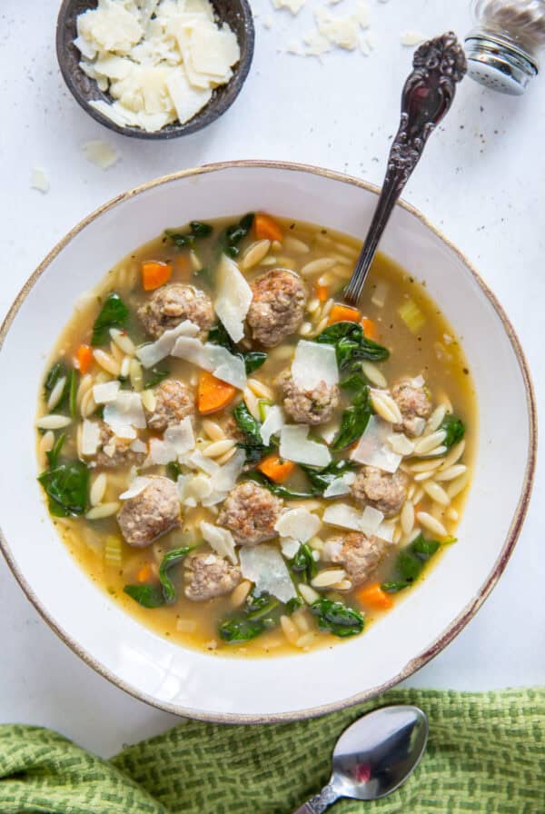 A bowl of soup with visible mini meatballs, spinach, carrots, and pasta, garnished with Parmesan flakes and served with a spoon.