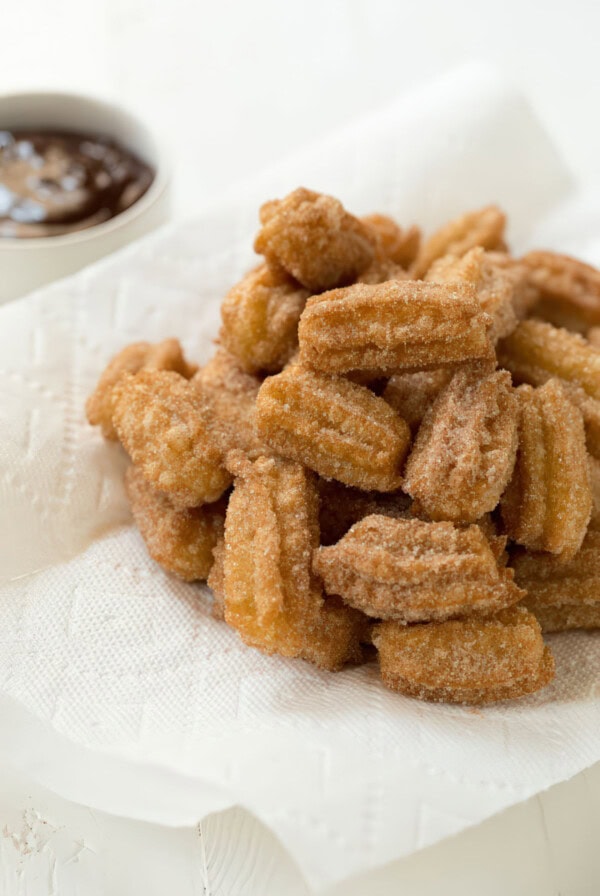 A heaping pile of freshly fried mini churros coated in cinnamon sugar, resting on a white paper towel with a bowl of chocolate sauce in the corner.