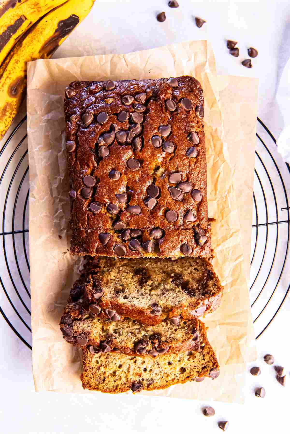 Overhead view of a partially sliced loaf of chocolate chip banana bread on parchment paper over a wire cooling rack, with ripe bananas and scattered chocolate chips nearby.
