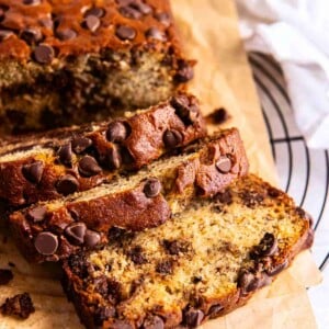 Sliced chocolate chip banana bread with a glossy, chip-studded crust and soft banana crumb, set on parchment paper near a wire cooling rack.