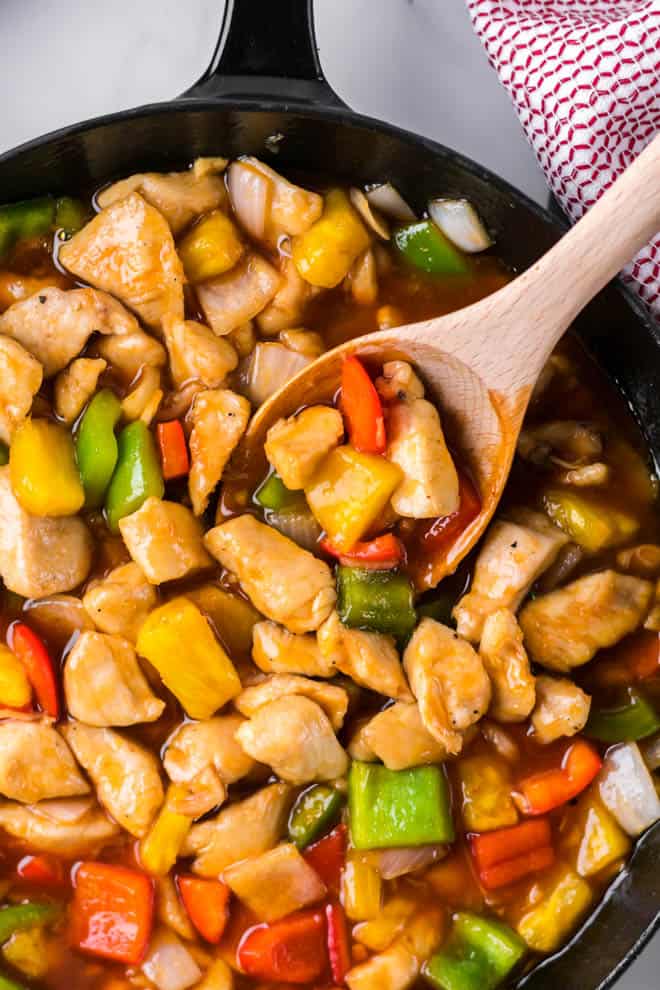 chicken with pineapple, bell peppers and sweet and sour sauce in a dark skillet with a wooden spoon