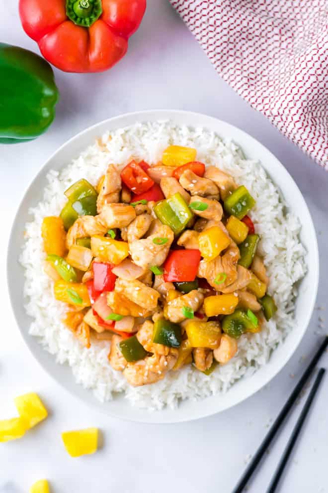 White plate filled with white rice and topped with sweet and sour chicken sitting on a table next to chop stix