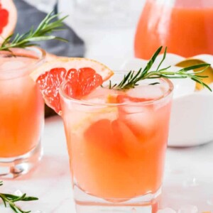 One clear glass with grapefruit gin fizz drink, ice cubes and a sprig of fresh rosemary sitting on a white table