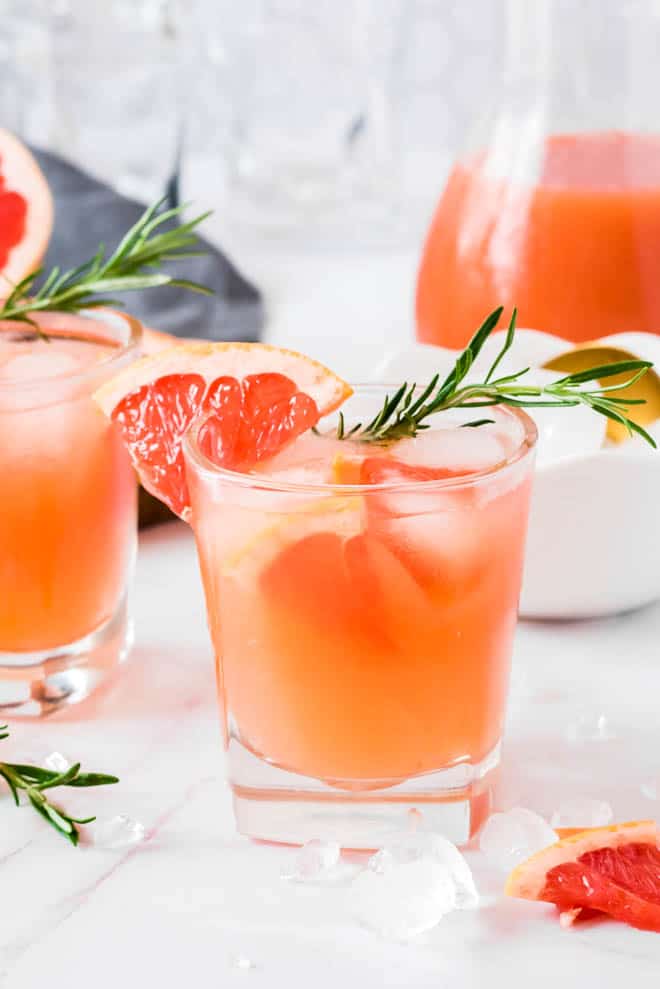 One clear glass with grapefruit gin fizz drink, ice cubes and a sprig of fresh rosemary sitting on a white table.
