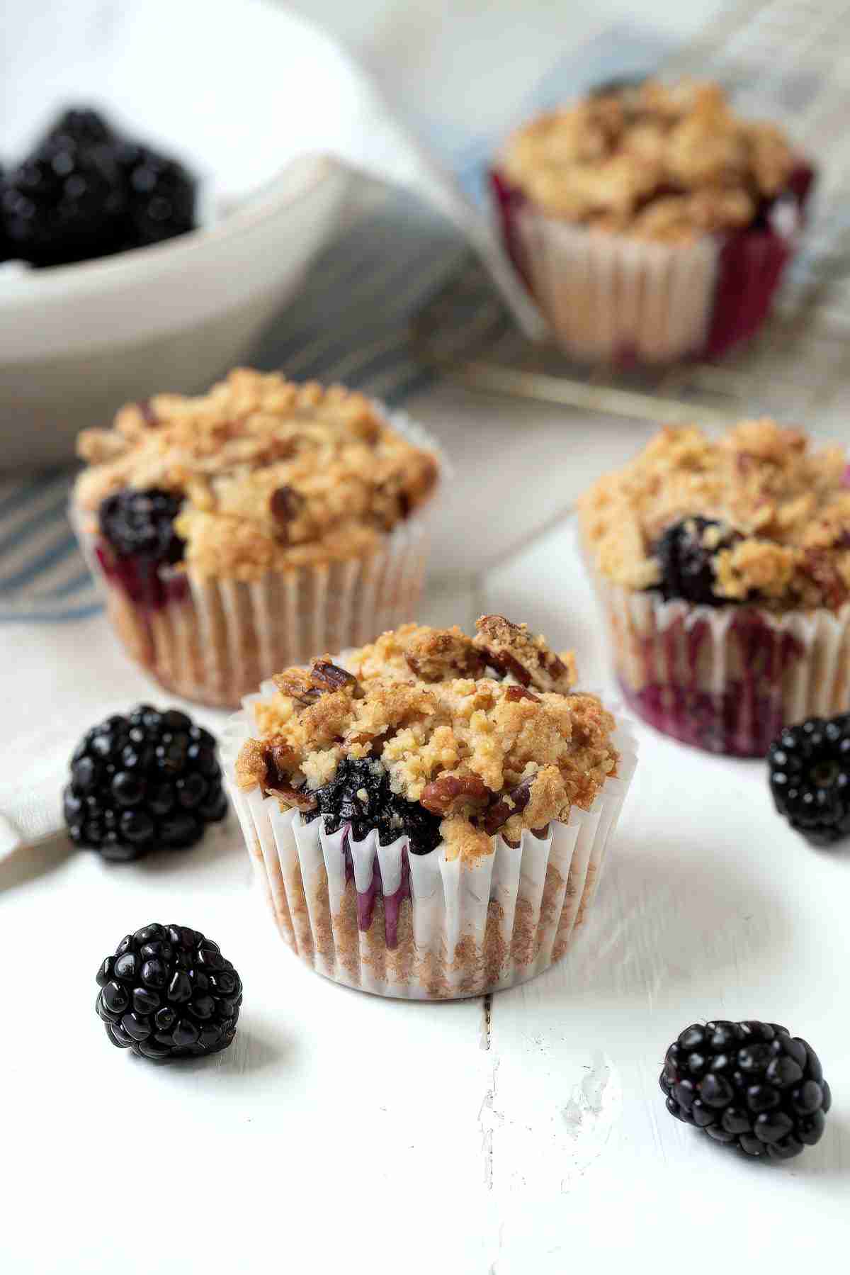 Blackberry muffins with crumble topping on a white surface with fresh blackberries scattered around