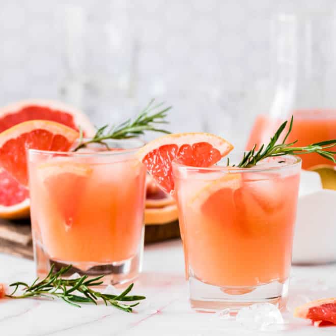 two glasses of grapefruit gin fizz sitting on a white table with grapefruit and rosemary sprigs on the side