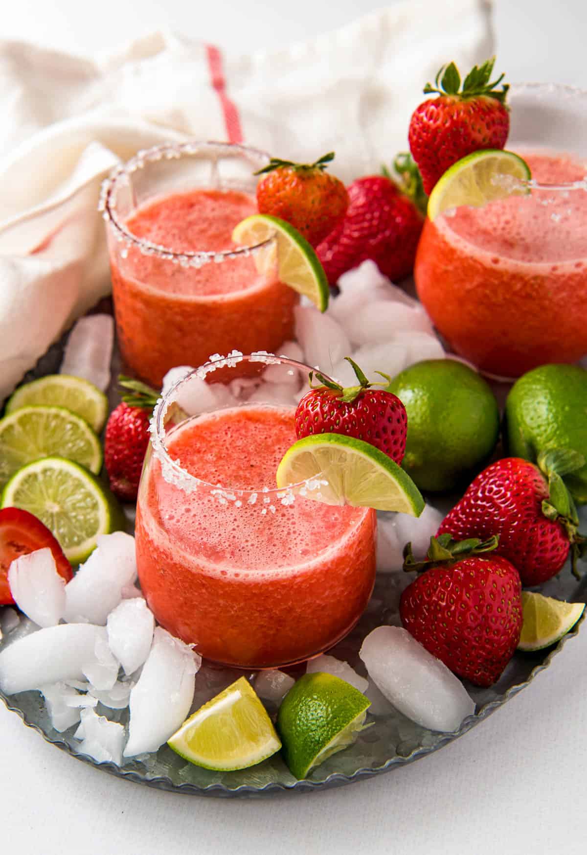 Three glasses of frozen strawberry margaritas on a serving tray garnished with ice cubs, strawberries and lime wedges.