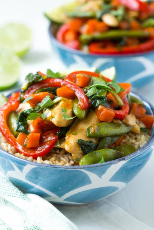 Close-up of a colorful chicken teriyaki stir fry served over brown rice in a blue patterned bowl, featuring bell peppers, carrots, snap peas, and fresh herbs.