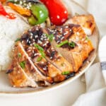 Close-up of sliced, charred chicken topped with sesame seeds and green onions, served alongside rice and roasted bell peppers