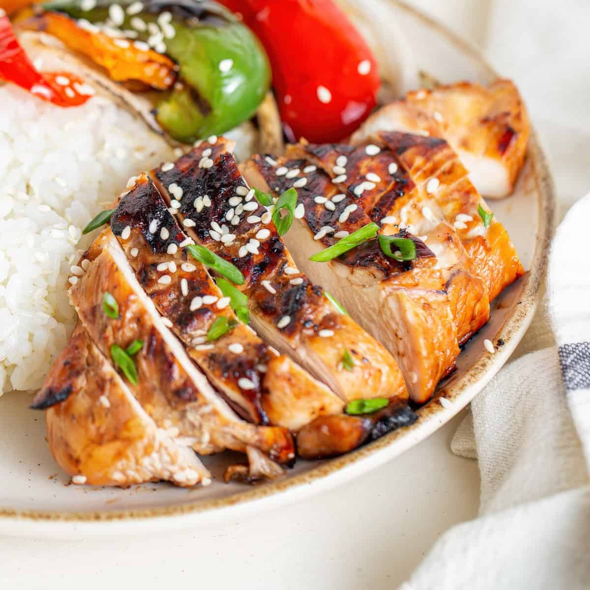 Close-up of sliced, charred chicken topped with sesame seeds and green onions, served alongside rice and roasted bell peppers