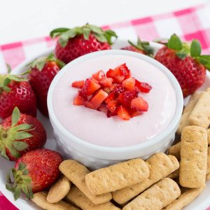 a bowl filled with strawberry cheesecake dip sitting on a plate with graham crackers and whole strawberries