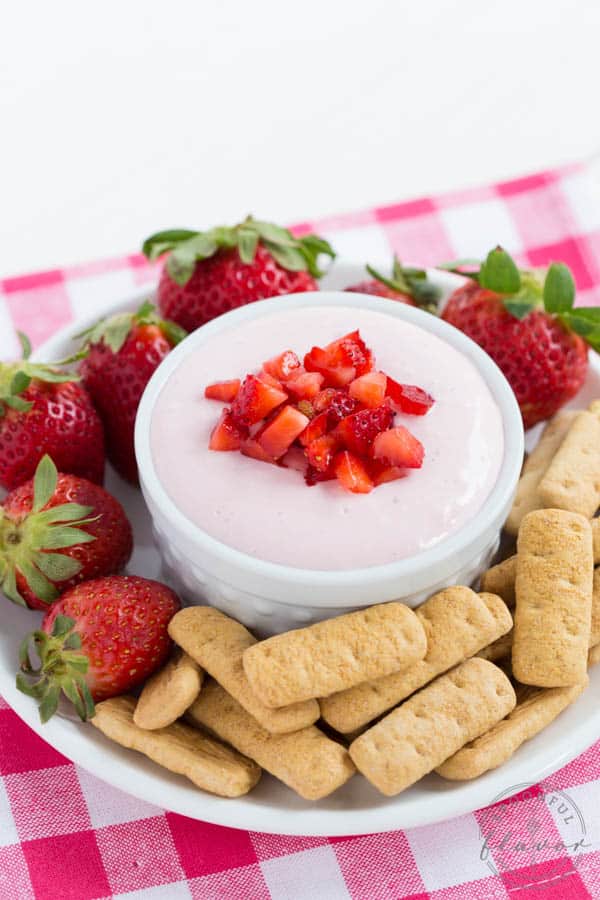 a bowl filled with strawberry cheesecake dip sitting on a plate with graham crackers and whole strawberries