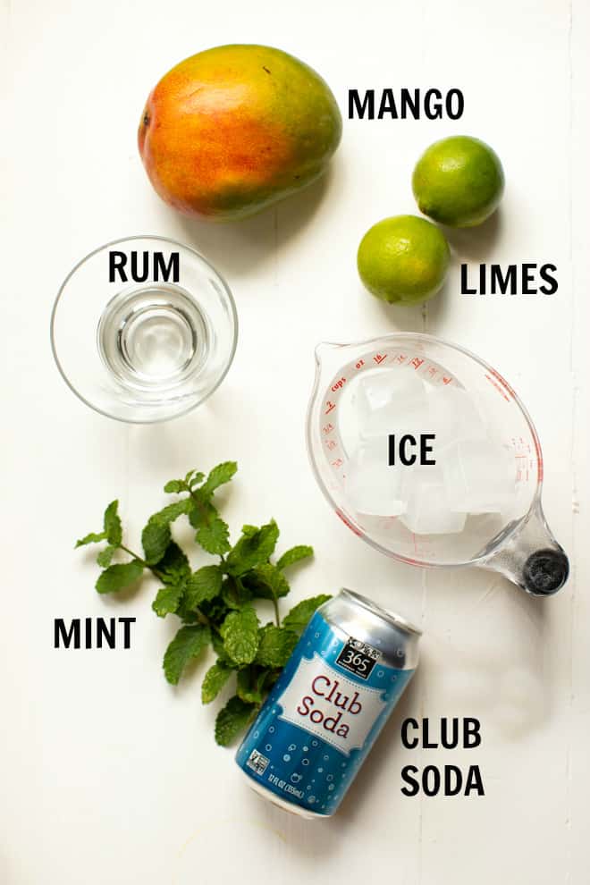 ingredients for mango mojito including mango, two limes, rum, ice, mint and club soda on a white table