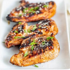 three pieces of grilled teriyaki chicken sitting on a white plate and garnished with sesame seeds and chives