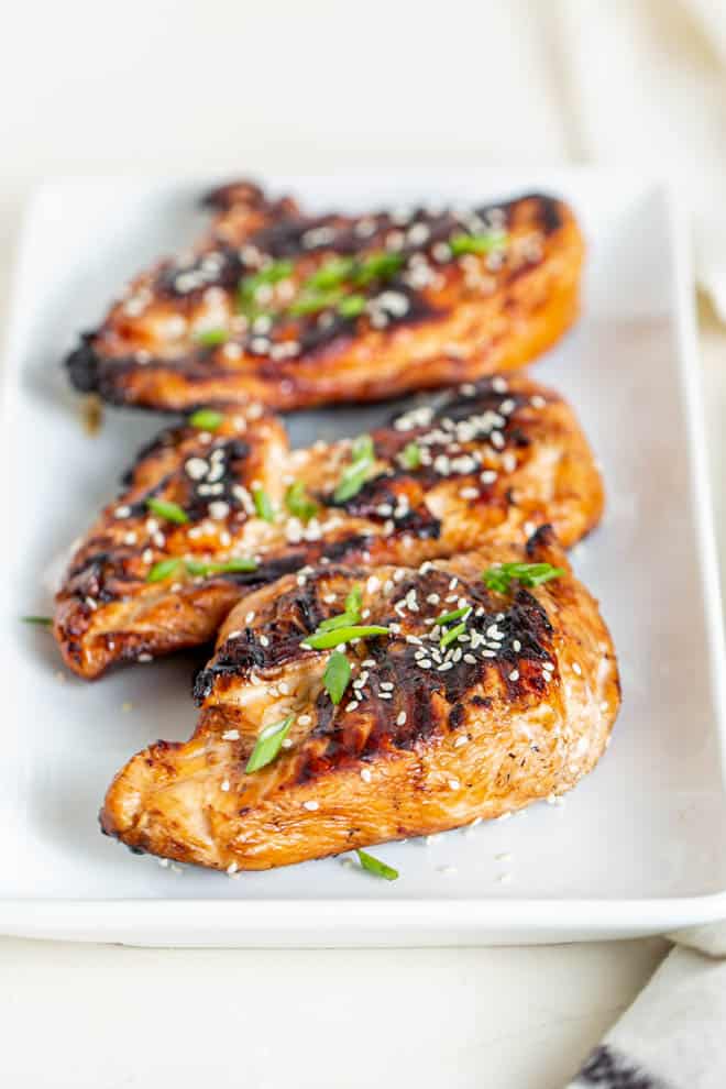 three pieces of grilled teriyaki chicken sitting on a white plate and garnished with sesame seeds and chives