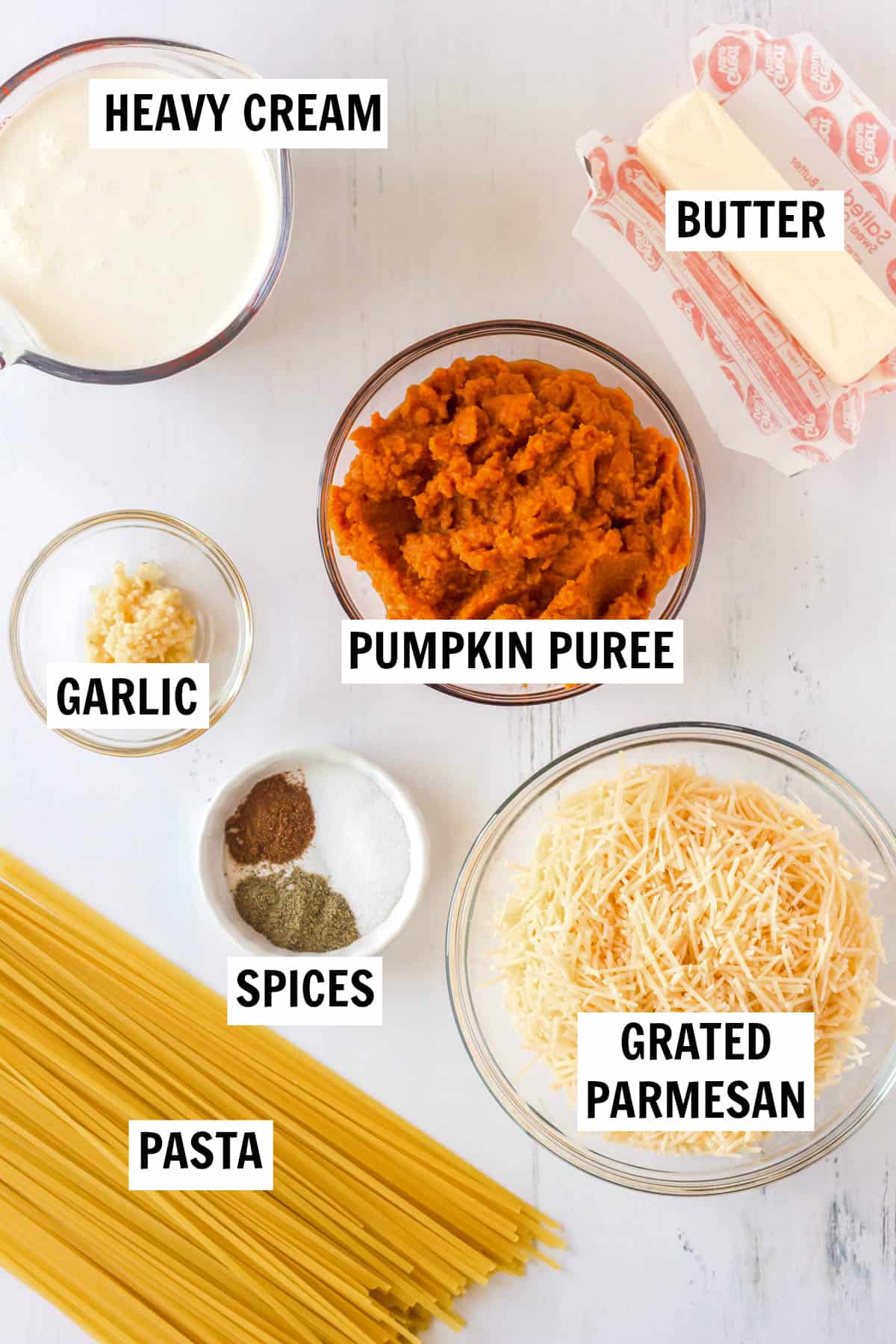 Ingredients for pumpkin pasta sauce sitting on a white countertop including pasta, pumpkin puree, grated parmesan, spices, garlic, heavy cream and butter. 