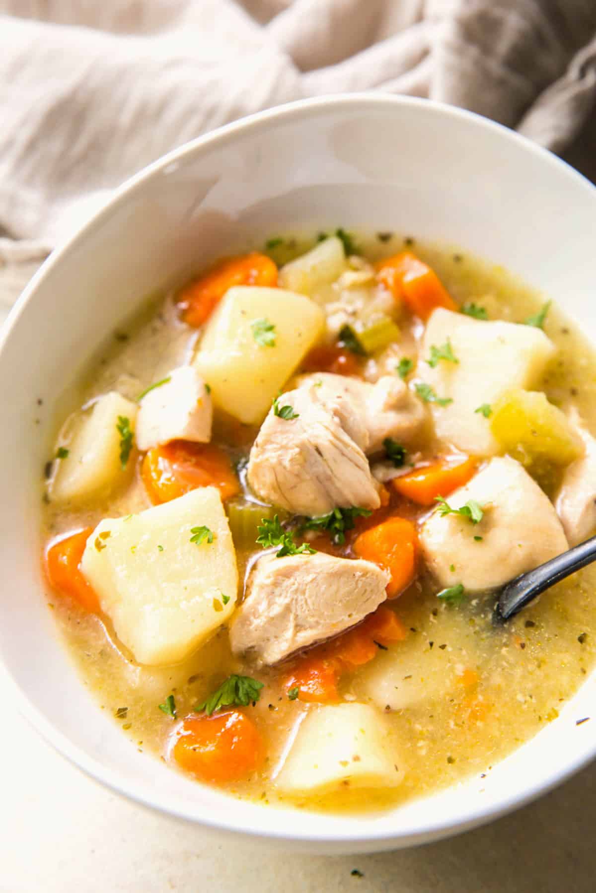 Close-up of chicken stew in a white bowl showing chunks of chicken, potatoes, carrots, and celery with parsley garnish.